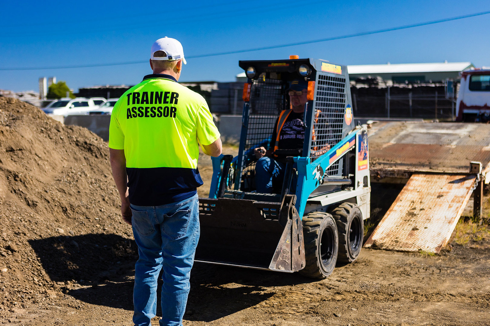 A Western Trainers training assessor wears a labeled high-viz shirt. He is instructing a learning driving a small skid-steer loader