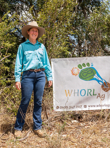 Marcia stands in front of a Whorl Ag sign hanging from a wire fence. The fence is in a brown paddock against bushy trees.