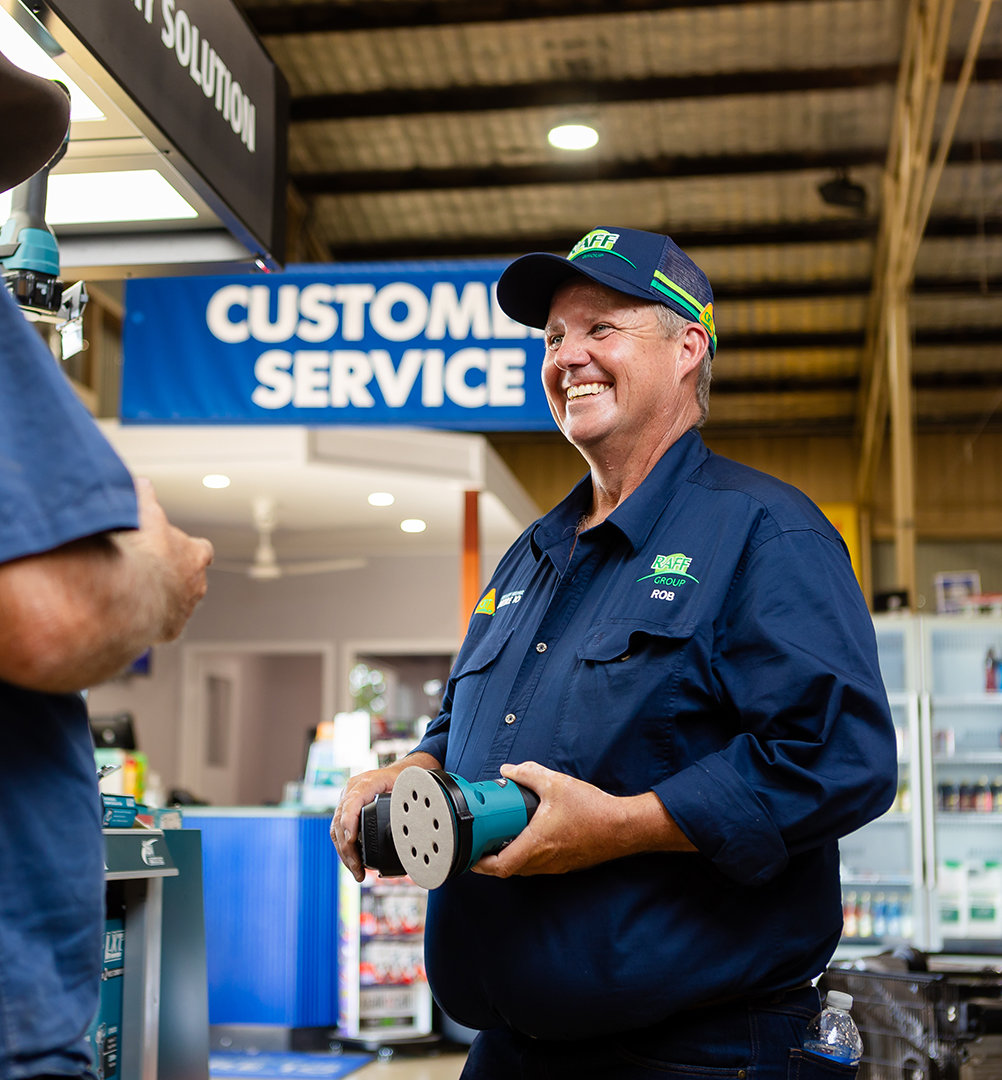 A RAFF Group employee holds an electric sander while talking to a customer. In the background you can see a 'customer service' banner.